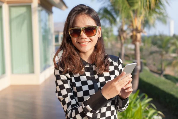 年轻的亚裔女子戴太阳眼镜和智能手机在手到 — 图库照片