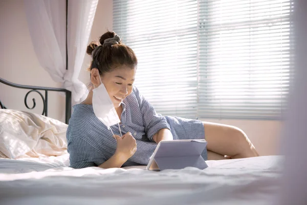 woman with protection mask in hand lying on sofa bed in home living room chatting to internet device