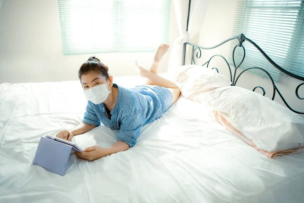 quarantine woman wearing protection mask lying on bed and chatting on internet device