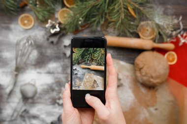 Kadın eli telefon tutuyor ve yemek pişirirken Noel kurabiyelerinin fotoğrafını çekiyor. Üst manzara, düz yatış. Noel havası, kış tatili modeli.