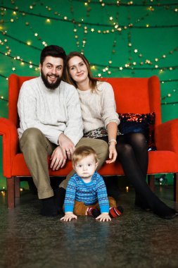Noel arifesindeki dost canlısı aile portresi: genç ebeveynler ve kameraya bakan küçük oğul. Noel ailesi görünüşü. Kırmızı kanepe, bahçeli yeşil duvar.