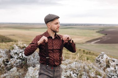 Tepenin tepesinde oturan, gömlek, şapka ve pantolon askısı takmış gülümseyen bir adamın portresi. Bir adam pantolon askısı çeker. Arka planda güzel bir manzara. Pantolon askısı reklamları