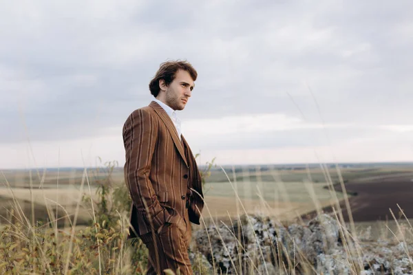 Pensive man in elegant suit looking away, hands in pockets. Standing on outdoor on the top of hill. Fields and hill background. Man thinking about future chalenges.