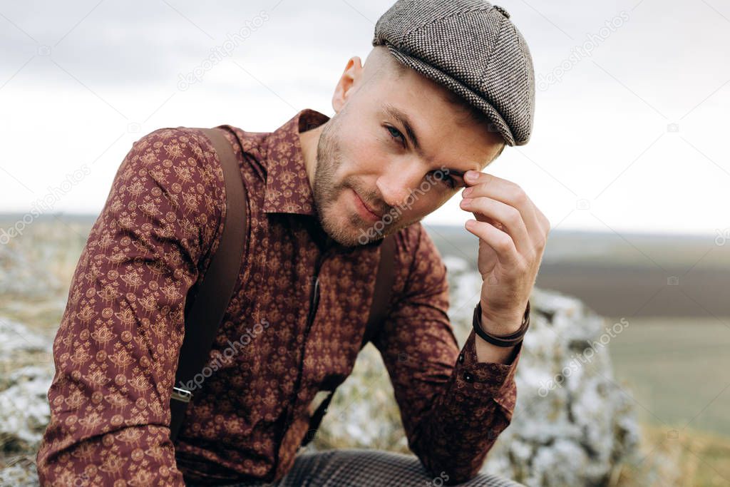 Portrait of pensive man in shirt, cap and suspenders, who sits on the rock top of the hill. Man is looking on camera. Beatuful landscape on background