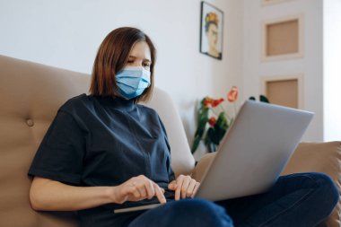 Evde dizüstü bilgisayarla gezen bir kız. Yüzünde koruyucu maske var. Sosyal izolasyon, karantina, sosyal uzaklık, evde kalma konsepti. Coronavirüs salgını.