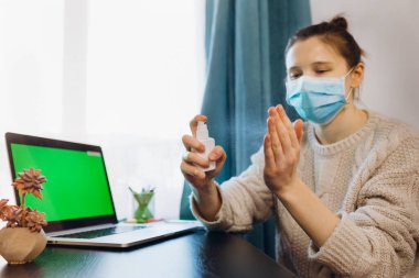 Koruyucu maske takan bir kadın evde çalışıyor. Koronavirüsü veya grip hastalığını önlemek için elleri antiseptik sprey kullanarak dezenfekte ediyor. Kişisel temizlik, karantina, evde kalma konsepti..