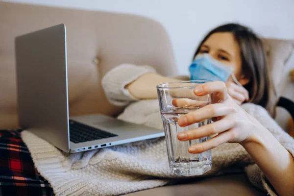 Woman lying on the sofa and drinking water. She has virus, wears protective mask not to infect other people. Social isolation, quarantine, stay home concept.Coronavirus pandemic