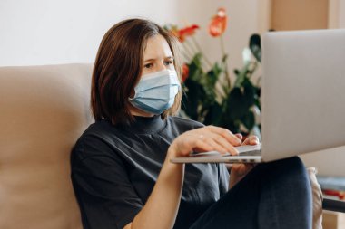 Evde dizüstü bilgisayarla gezen, online eğitim gören bir kız. Yüzünde koruyucu maske var. Sosyal izolasyon, karantina, yaşam tarzı, ev konsepti. Coronavirüs salgını