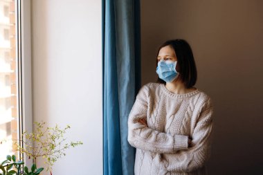 Coronavirus salgınları ve grip salgını sırasında virüslerden, mikroplardan ve bakterilerden korunmak için tıbbi maske takan kadın portresi. Pencereye bakıyorum. Sosyal izolasyon, karantina kavramı.