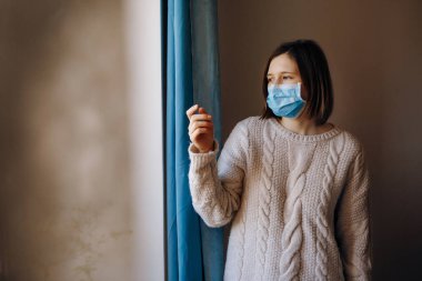 Coronavirus salgınları ve grip salgını sırasında virüslerden, mikroplardan ve bakterilerden korunmak için tıbbi maske takan kadın portresi. Pencereye bakıyorum. Sosyal izolasyon, karantina kavramı.