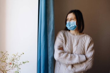 Coronavirus salgınları ve grip salgını sırasında virüslerden, mikroplardan ve bakterilerden korunmak için tıbbi maske takan kadın portresi. Pencereye bakıyorum. Sosyal izolasyon, karantina kavramı.