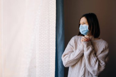 Coronavirus salgınları ve grip salgını sırasında virüslerden, mikroplardan ve bakterilerden korunmak için tıbbi maske takan kadın portresi. Pencereye bakıyorum. Sosyal izolasyon, karantina kavramı.