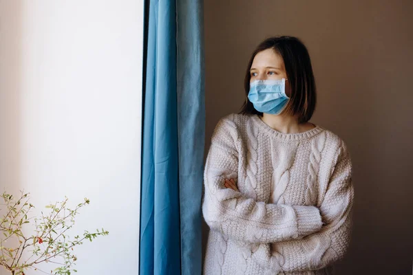 Coronavirus salgınları ve grip salgını sırasında virüslerden, mikroplardan ve bakterilerden korunmak için tıbbi maske takan kadın portresi. Pencereye bakıyorum. Sosyal izolasyon, karantina kavramı.