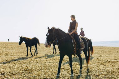 Arkadan at biniyor, geniş bir araziye ve ormana bakıyor. Dostlar at kulübünde boş vakit geçirmenin keyfini çıkarıyorlar. Hobi, spor, tatil köyü ve çiftlik konsepti.