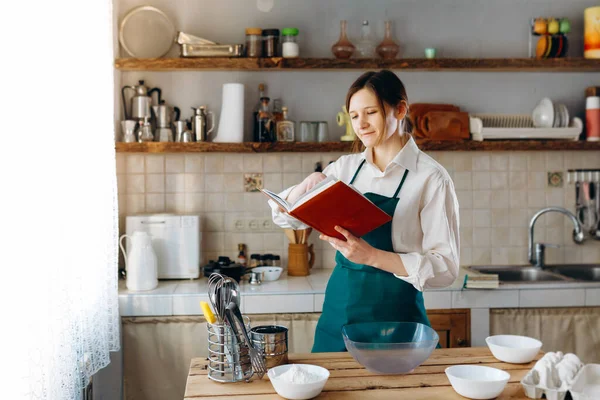 Young woman standing in the middle of the kitchen with recipe book in her hands. She looking in notebook and trying to choose what to cook. Cooking at home concept, lifestyle.