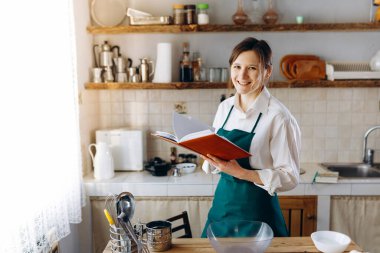 Mutfağın ortasında elinde yemek tarifi kitabıyla duran genç bir kadın. Kameraya bakıyor ve gülümsüyor. Ev konseptinde aşçılık, yaşam tarzı.