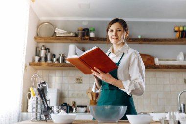 Mutfağın ortasında elinde yemek tarifi kitabıyla duran genç bir kadın. Kameraya bakıyor ve gülümsüyor. Ev konseptinde aşçılık, yaşam tarzı.