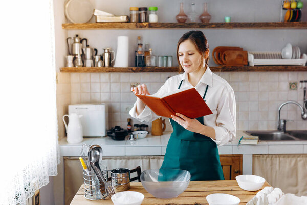 Young woman standing in the middle of the kitchen with recipe book in her hands. She looking in notebook and trying to choose what to cook. Cooking at home concept, lifestyle. 