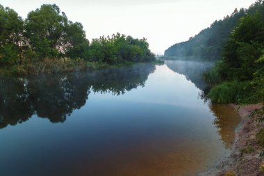 Bir orman nehrinin kıyısındaki sarp bir uçurum mavi bir gökyüzüne karşı bir çayır boyunca akıyor..