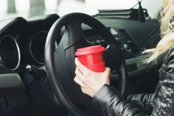 Mujer con una taza roja de bebida caliente mantiene la rueda de un coche — Foto de Stock