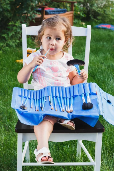 Girl sits on the chair in the garden with a big collection of brushes for make up