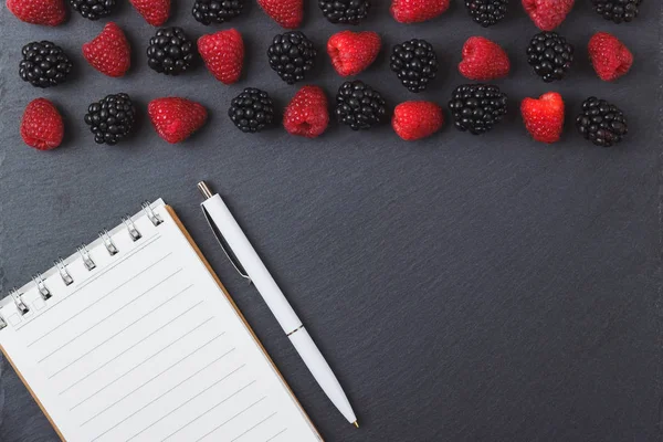 Red raspberries, blackberry, notebook and a pen on the black slate stone background — Stock Photo, Image