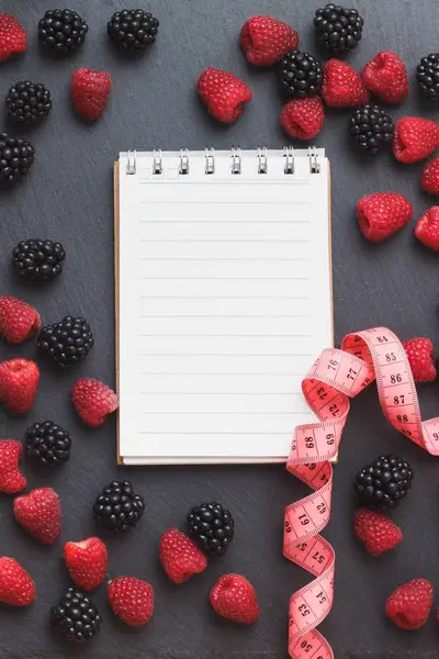 Red raspberries, tape measure, notebook and a pen on the black slate stone background — Stock Photo, Image