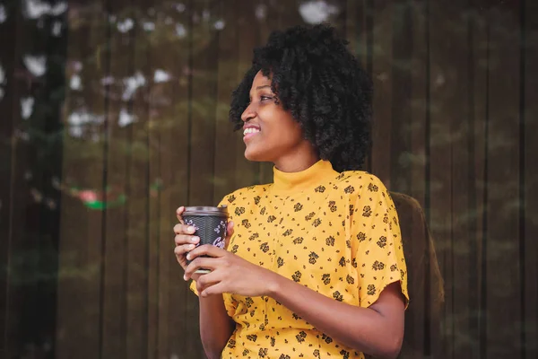 Young dark-skinned woman dressed in casual clothes holding cup of hot drink