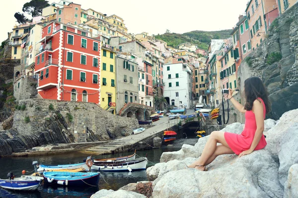 Vista trasera de una chica vestida de rojo sentada en las piedras como una sirena tomando fotos con una cámara sin espejo del paisaje italiano de la Riviera, Riomaggiore, Cinque Terre, Italia. Filtro Vintage — Foto de Stock