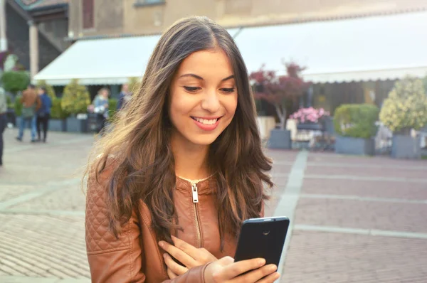 Счастливая молодая женщина в коричневой куртке читает сообщение на смартфоне — стоковое фото