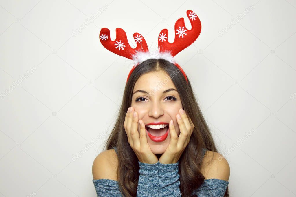 Surprised excited christmas woman on gray background. Beautiful happy christmas girl with reindeer horns on her head holding her face. Cheerful female model joyful. 