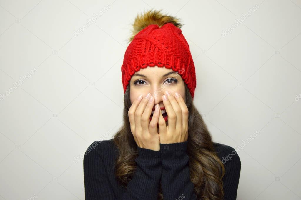 Happy shy girl with pompon hat hide her mouth with hands on gray background
