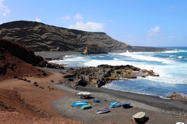 Tekneler ile El Golfo Koyu'nun nefes kesen manzarası: kırmızı zemin, mavi deniz ve arka plan, Lanzarote, Kanarya Adaları volkanik Dağları ile siyah kum