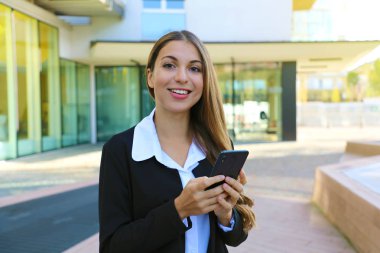 Genç iş kadını dışarıda kameraya bakarak akıllı telefon kullanıyor.