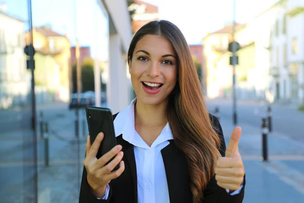 Bonita jovem empresária segurando telefone inteligente dá polegar na rua . — Fotografia de Stock