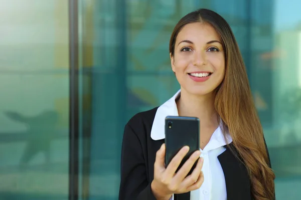 Mulher de negócios feliz jovem verificando seu telefone inteligente e olhando para a câmera fora do escritório. Espaço de cópia . — Fotografia de Stock
