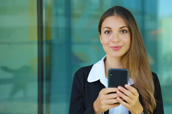 Mulher de negócios jovem verificando seu telefone inteligente e olhando para a câmera fora do escritório. Espaço de cópia . — Fotografia de Stock