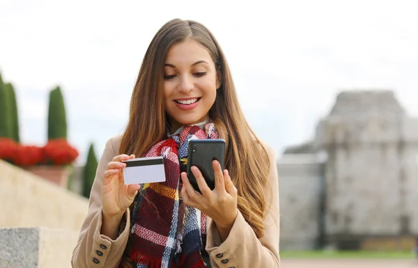Young shopper woman holding credit card buying online with smart phone sitting outdoor.