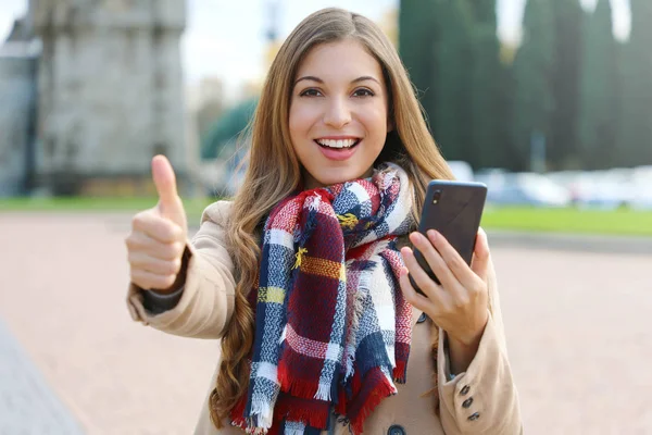 Mujer de moda joven bastante sonriente usando teléfonos inteligentes muestra el pulgar hacia arriba al aire libre en invierno . —  Fotos de Stock
