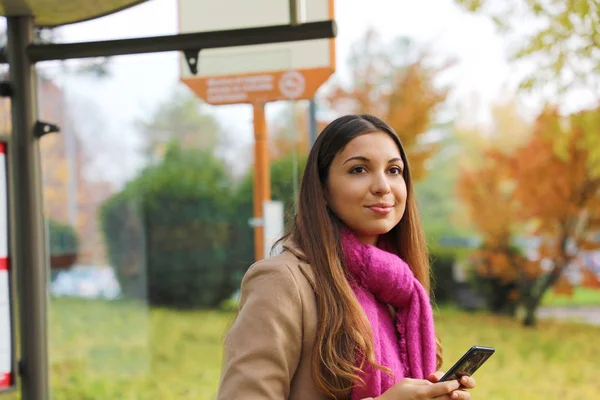 Personas y transporte público. Hermosa joven sosteniendo el teléfono móvil esperando el autobús en la parada de autobús . — Foto de Stock