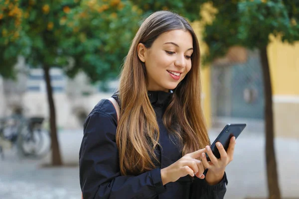 Retrato de una hermosa mujer sonriente usando un teléfono móvil en la calle de la ciudad con árboles de mandarinas en el fondo . — Foto de Stock
