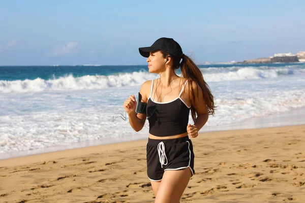 Running woman with black cap listening to music on her phone sports armband with touchscreen and earphones on summer beach