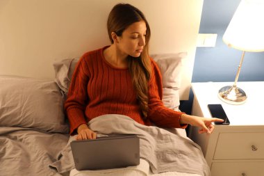 Student girl working on laptop on bed distracted by phone at the night time clipart