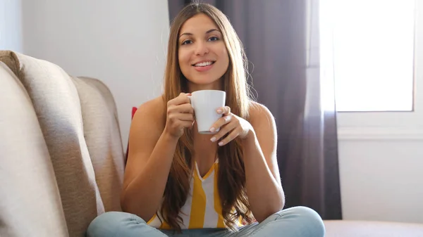 Šťastná studentka sedí na pohovce se zkříženýma nohama a drží hrnek čaje nebo cappuccino doma v obývacím pokoji a dívá se do kamery — Stock fotografie
