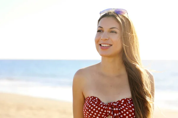 Közelkép portré vonzó mosolygós lány hosszú haj pózol a strandon. Divat bikinit és napszemüveget hord a kezén, oldalra néz.. — Stock Fotó