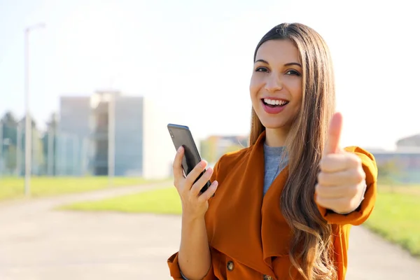 Joven hermosa mujer casual sosteniendo el teléfono inteligente que muestra el pulgar hacia arriba en la calle de la ciudad en primavera. Copiar espacio . —  Fotos de Stock