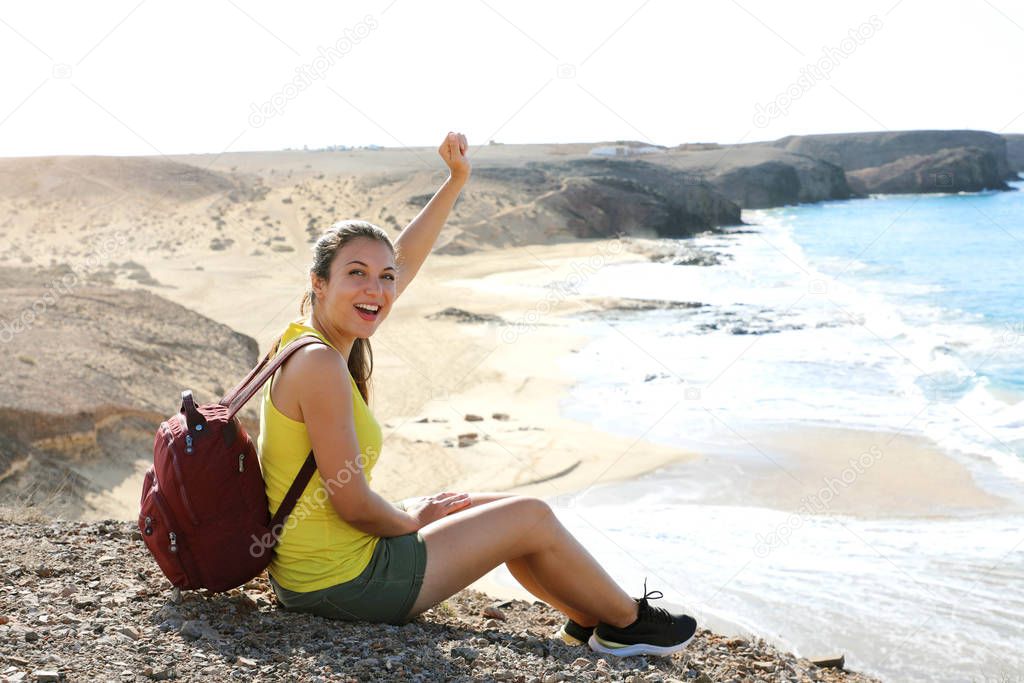 Portrait of young female hiker reaches top of mountain and celebrates with amazing landscape on the background. Happy success backpacker girl celebrating outdoor raising her fist up in exultation.