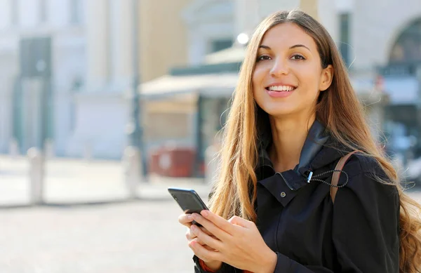Счастливая Женщина Улыбается Ходит Улице Используя Смартфон Глядя Камеру — стоковое фото