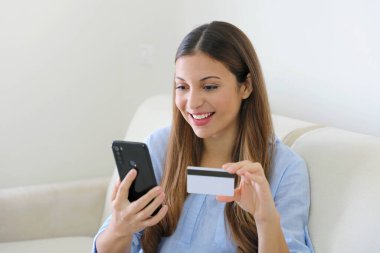 İnternetten kredi kartı ve akıllı bir telefonla satın alan bir kadın evindeki oturma odasında rahat bir şekilde oturuyor.