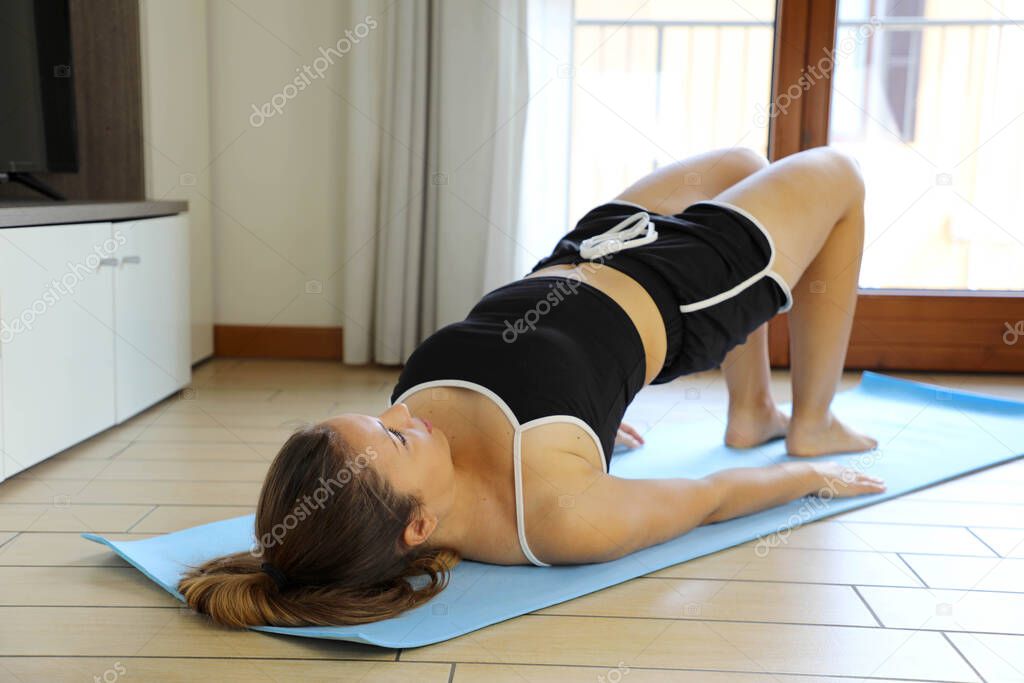 Glute Bridge. Fitness young sporty woman doing the half bridge pose at home. Gymnastics practices yoga warming up exercises for spine, backbend, strengthening back and shoulders muscles.